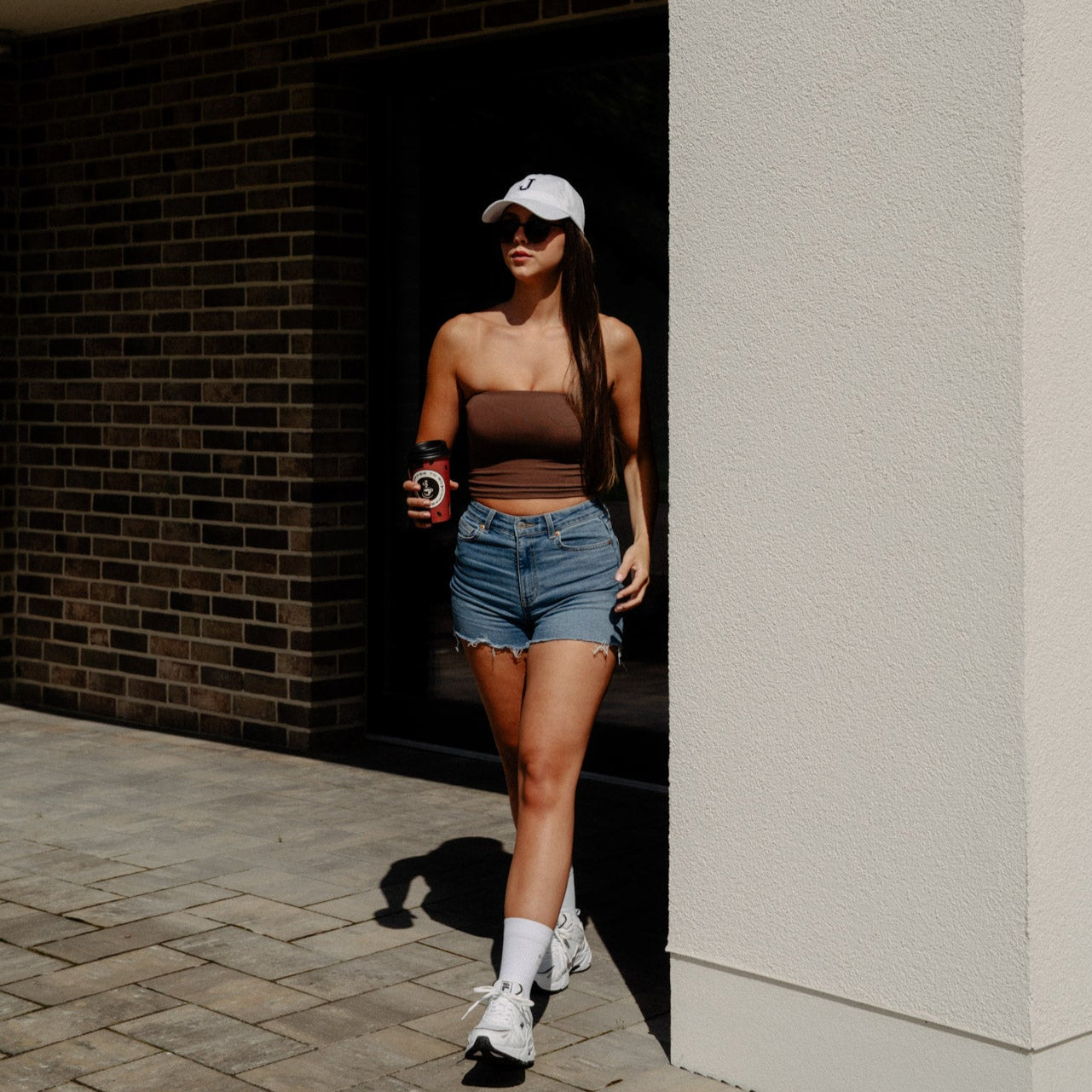 Eine Frau läuft aus einer modernen Wohnung in Freizeitkleidung und einem Cafe in der Hand aus dem Haus. Dazu trägt sie die Sportsocken und Sportschuhe.